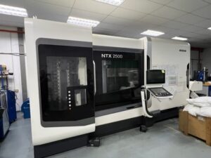 A white NTX 2500 CNC machine with a control panel is situated in an industrial workshop setting under fluorescent lighting.