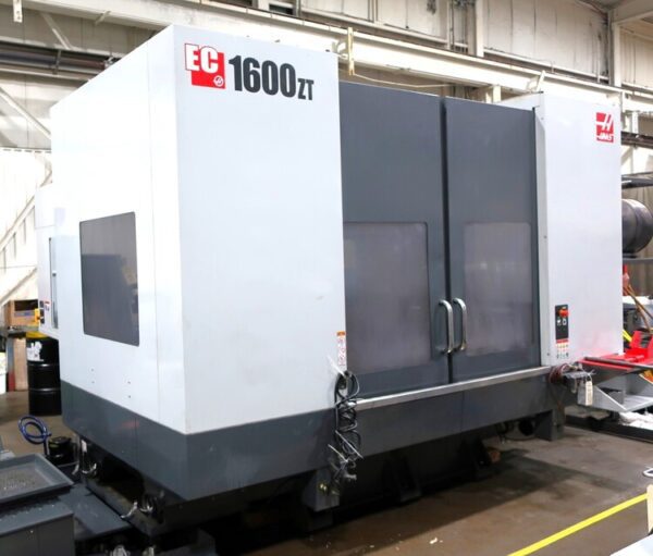 A large industrial CNC machine labeled EC 1600ZT, with a white exterior and dark gray panel doors, is positioned in a factory setting.