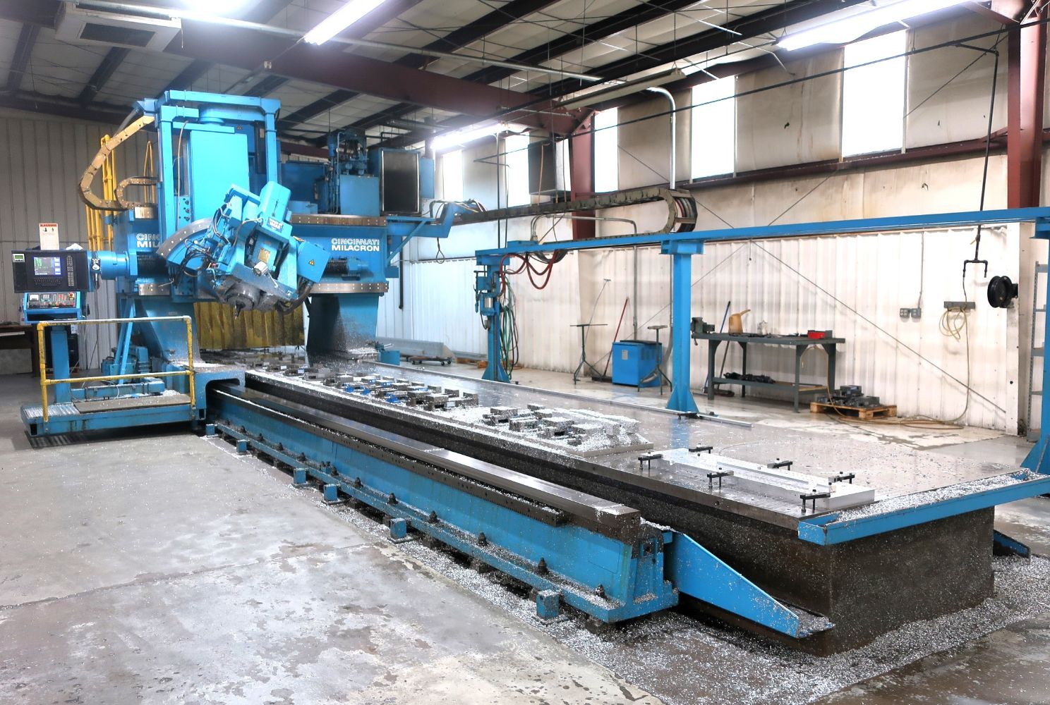 Large blue industrial CNC machine in a workshop, surrounded by metal shavings on the floor. Concrete walls and metal beams are visible in the background.