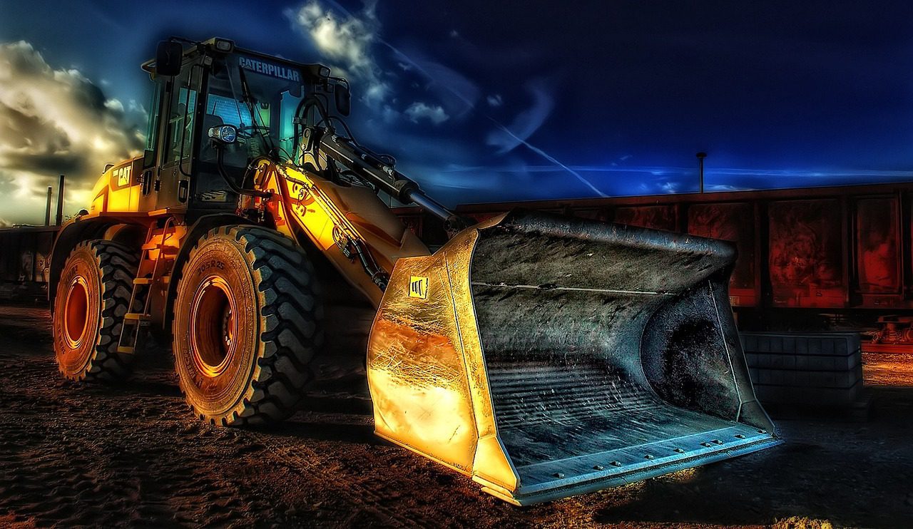 A large yellow bulldozer with a front bucket is parked on a dirt surface against a dramatic sky backdrop.