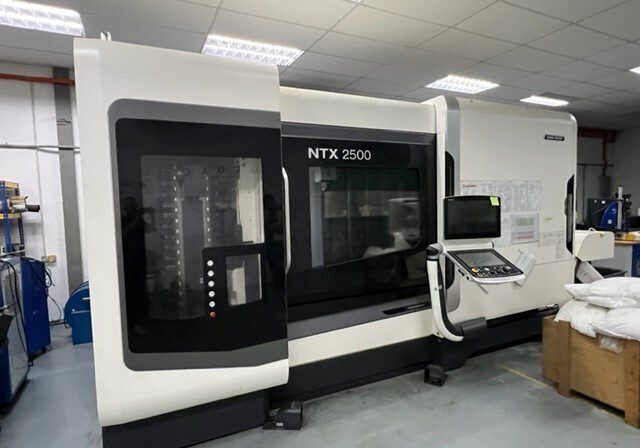 A white NTX 2500 CNC machine with a control panel is situated in an industrial workshop setting under fluorescent lighting.