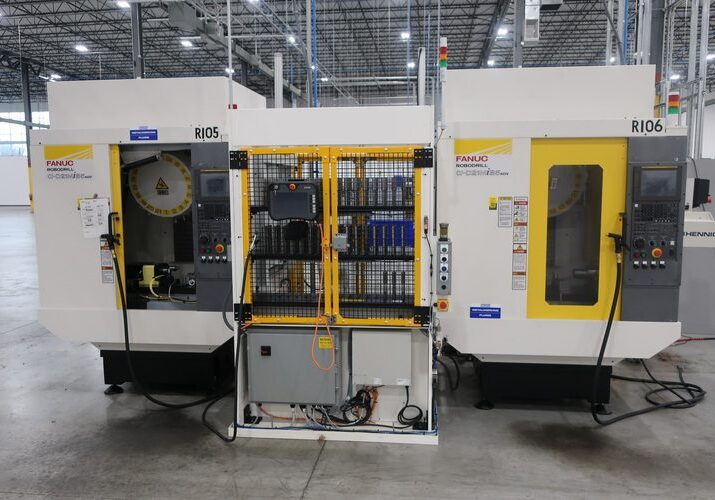 Two industrial CNC machines sit side by side in a spacious factory setting. They have control panels and safety enclosures with yellow doors.